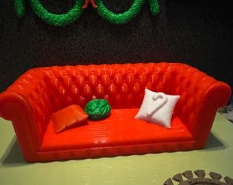 May include: A miniature red sofa with button tufting, two pillows, and a green decorative item. One pillow is orange, and the other is white with a curved design. A green garland hangs above the sofa.