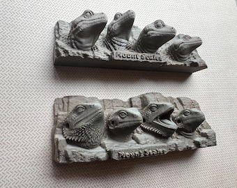May include: Two gray sculptures of lizard heads emerging from a rock formation, resembling Mount Rushmore. The text "Mount Scalie" is inscribed on the base of each sculpture. The sculptures are displayed on a light patterned surface.