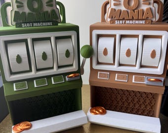 May include: Two plastic slot machine toys labeled BUG O MANIA. One is olive green with white reels and green leaf icons, the other is tan with white reels and tan leaf icons. Both feature a side lever, a coin slot, and metallic orange coins.