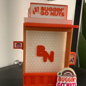 May include: A 3D printed model of a store front with the words "Buggin' Go Nuts" on the sign. The store is orange and white with a "BN" logo on the front. There is a sticker on the bottom right corner that says "Buggin' Go Nuts" with the text "America Runs From Nuts".