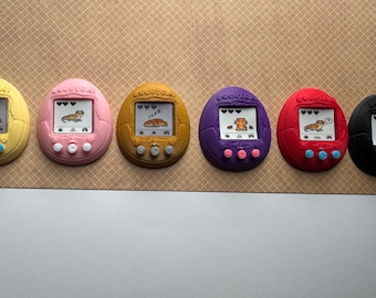 May include: Six colorful, egg-shaped digital pet toys. Each has a small screen displaying a cartoon animal, with buttons below. Colors include yellow, pink, gold, purple, red, and black. The screens show a sleeping animal or a frog.