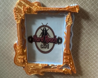 May include: A rectangular picture frame with a metallic gold finish, featuring a border constructed from stylized electric guitar shapes. The center insert displays a circular logo with a cockroach illustration and the text Hard Roach CAFE.
