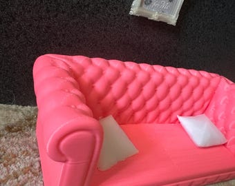 May include: A bright pink miniature sofa with a tufted back and two white square pillows. A framed picture with the text "Diamonds for Her, Bugs for Me" hangs above the sofa.