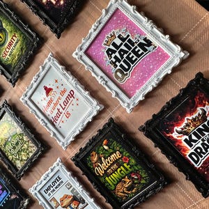 May include: A selection of framed art prints with diverse designs and text. The frames are black and white with decorative detailing. Prints feature phrases such as "All Hail Queen", "Welcome to the Jungle", and "King of Dragons".