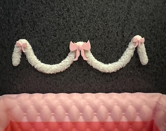 May include: A decorative garland with a fluffy white texture, adorned with three pink bows. The garland is suspended above a pink, quilted surface, creating a soft and feminine aesthetic. The background is a dark gray.