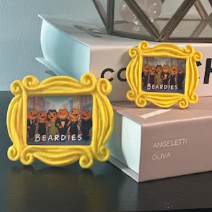 May include: Two yellow picture frames with a cartoon image of frogs in suits and the word "BEARDIES". The frames are in a decorative style, and are placed on top of books.