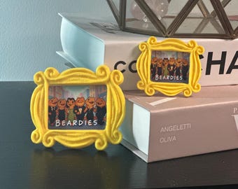 May include: Two yellow picture frames with a cartoon image of frogs in suits and the word "BEARDIES". The frames are in a decorative style, and are placed on top of books.