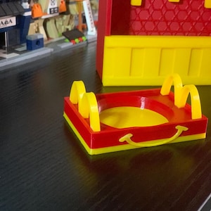 May include: A red and yellow plastic tray with a smiley face design. The tray has a raised edge and is shaped like a small food tray.