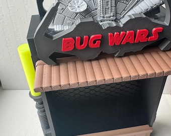 May include: A 3D printed display stand featuring a gray spaceship model at the top, a black frame with the red text "BUG WARS" in the center, a brown roof-like ledge, a black honeycomb-patterned back panel, and a bright neon yellow side accent.