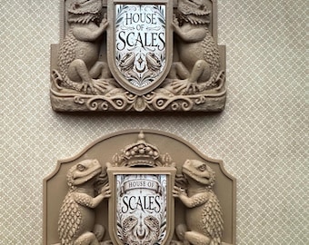 May include: Two beige wall plaques, each featuring a shield with the text "HOUSE OF SCALES" flanked by two lizard-like creatures. The plaques have a crown at the top and decorative scrollwork.