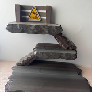 May include: A multi-tiered reptile habitat with a wooden frame and textured, gray-brown platforms. A yellow triangle sign with a lizard graphic and the words "Reptile Zone" is attached to the top. The design mimics a natural rock formation.