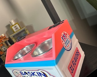 May include: A colorful, custom-made ice cream container with the text "Baskin in Bugs" and "31 Flavors." The container is white, pink, and blue, with two silver metal bowls. The design is reminiscent of an ice cream shop.