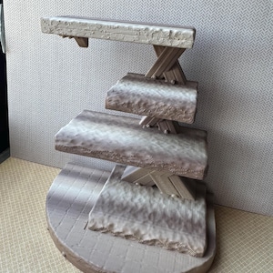 May include: A multi-tiered, decorative display stand with a stone-like appearance. The stand features a semi-circular base, three rectangular shelves, and an ornate top shelf. The shelves are supported by an X-shaped structure. The color is a blend of gray and brown.