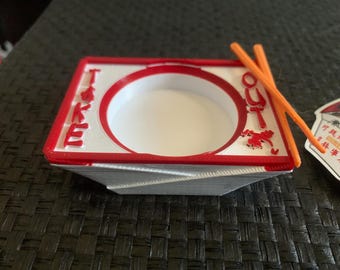 May include: A red and white takeout container with the words "Take Out" printed on the side. Two orange chopsticks are placed on top of the container.