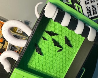 May include: A green and black box with a striped top and the words "BEETLE JUICE" on a sign. The box has a green interior with a diamond pattern and black bat cutouts. A white, curved handle is attached.