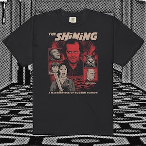 May include: Black t-shirt featuring a graphic from the movie "The Shining." The design includes the film's title, images of characters, and the tagline "A Masterpiece of Modern Horror."