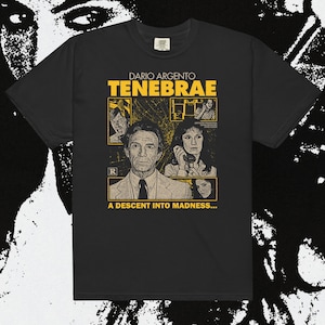 May include: Black t-shirt featuring a movie poster design for Dario Argento's "Tenebrae." The design includes the film's title, character portraits, and the tagline "A Descent Into Madness..." in yellow and tan.
