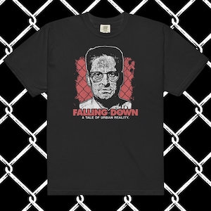 May include: A black short-sleeve t-shirt featuring a graphic print of a man wearing glasses. Behind the portrait is a red chain-link fence pattern. Below the image, the text reads FALLING DOWN A TALE OF URBAN REALITY in red and white lettering.