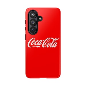 Puede incluir: Una funda de smartphone de color rojo brillante con el logotipo blanco de Coca-Cola impreso en el centro. La funda tiene un módulo de cámara negro que alberga tres lentes circulares y un pequeño flash, situado en la esquina superior izquierda.