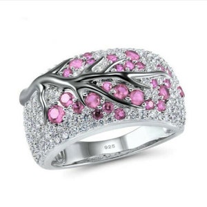 May include: A silver ring with a floral design. The ring features a band covered in small, clear stones, with a branch-like design in the center adorned with pink gemstones. The ring is marked with "925".