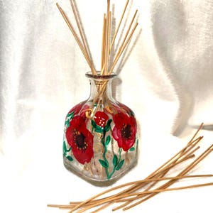 May include: A clear glass reed diffuser bottle with a square base, hand-painted with vibrant red poppy flowers, green stems, and gold swirl accents. The bottle contains several thin wooden reeds, with additional loose reeds placed nearby.