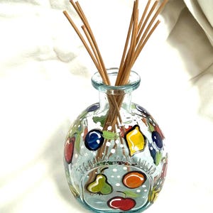 May include: A clear glass reed diffuser bottle with a rounded shape, featuring hand-painted colorful fruit motifs including apples, pears, and lemons, accented by white polka dots. Several thin, light brown wooden reeds extend from the narrow neck.