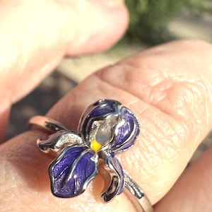 Puede incluir: Anillo de plata con un diseño detallado de flor de iris. La flor tiene pétalos morados vibrantes con un centro amarillo, engastados en una banda de plata. Una pieza de joyería.