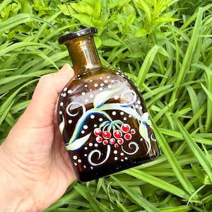 May include: A brown glass bottle with a black top, decorated with white dots, red berries, and blue and green leaves. The bottle is held against a backdrop of green foliage.