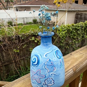 May include: A hand-painted blue vase with a coastal theme, featuring starfish, seashells, and swirls in white and pink. The vase holds blue and yellow dried flowers, set against a natural outdoor backdrop.