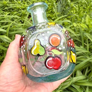 May include: A clear glass bottle with a painted fruit design. The bottle features a pear, cherry, and orange, with white polka dots and colorful accents. The bottle has a light blue rim and base.