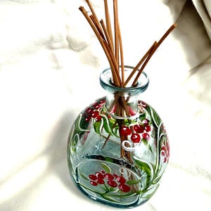 May include: A clear glass essential oil diffuser bottle with a painted design of red berries and green leaves. Brown reeds are inserted into the bottle. The bottle is round with a narrow neck.