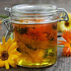 May include: A clear glass jar with a metal wire bail closure filled with golden yellow oil and submerged orange calendula flowers, surrounded by several bright orange and yellow calendula blossoms on a weathered wooden surface.