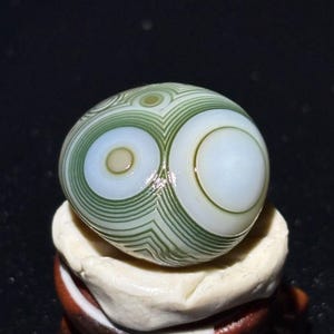 Può includere: Una pietra levigata di forma ovale con un motivo unico di cerchi e linee concentriche nei toni del verde, bianco e crema. La pietra è esposta su un piccolo supporto bianco sporco. Il design ricorda un occhio.