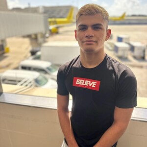 May include: A young man wearing a black t-shirt with a red box that says "BELIEVE" in white letters. He is standing in front of a window with a yellow airplane in the background.