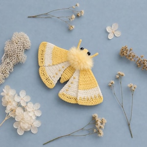 May include: A handcrafted crochet moth with yellow and white striped wings and a fluffy yellow body. The moth has black bead eyes and small antennae. The moth is surrounded by dried flowers on a pale blue background.