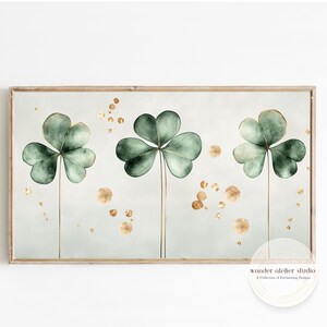 Könnte beinhalten: Ein gerahmter Druck mit einem Aquarelldesign aus drei grünen Kleeblättern mit goldenen Akzenten auf hellblauem Hintergrund. Der Druck hat einen weißen Rand und einen goldfarbenen Rahmen.