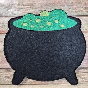 May include: A black cauldron-shaped decoration with a green, glittery potion bubbling inside. The cauldron has a textured surface and a black stitched border. The potion has lighter green bubbles. The decoration is on a wooden surface.