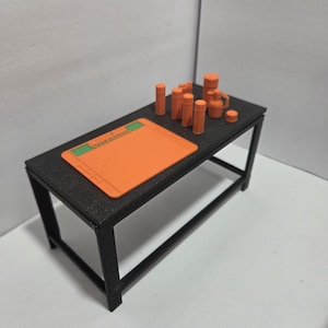 Peut inclure: Une petite table noire avec un tapis orange et plusieurs récipients cylindriques orange et un petit pichet. Le tapis porte du texte vert. La table est en métal noir.