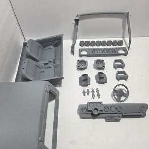 以下が含まれることがあります： ダッシュボード、ステアリングホイール、シート、ロールケージなど、グレーの3Dプリントされた車の内装部品のコレクション。これらのコンポーネントは白い表面に配置されており、モデルまたはDIYプロジェクトを示唆しています。