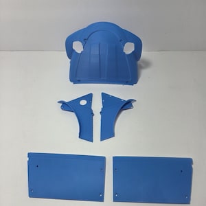 May include: Blue plastic car parts arranged on a white surface. The parts include a large, curved piece with two holes, two smaller curved pieces, and two rectangular panels. The pieces appear to be 3D printed.