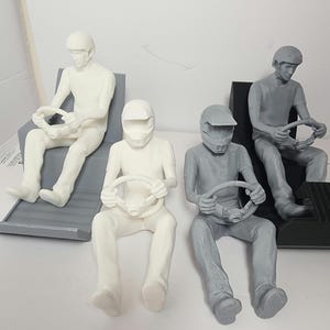 Puede incluir: Cuatro figuras en miniatura de pilotos de carreras sentados, cada uno sosteniendo un volante. Dos son de color gris claro y dos son blancos. Llevan cascos y están sentados en asientos de carreras. El fondo es blanco liso.
