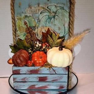May include: A seasonal autumn decoration. It features a wooden crate painted blue and red, filled with pumpkins, gourds, and foliage. The crate sits beneath a wooden sign with a pumpkin painting, framed with rope.