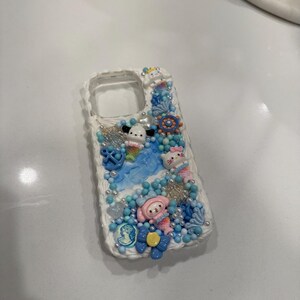 May include: A white phone case decorated with blue, pink and white 3D embellishments. The case features a mermaid theme with seashells, starfish and a small mermaid figurine.