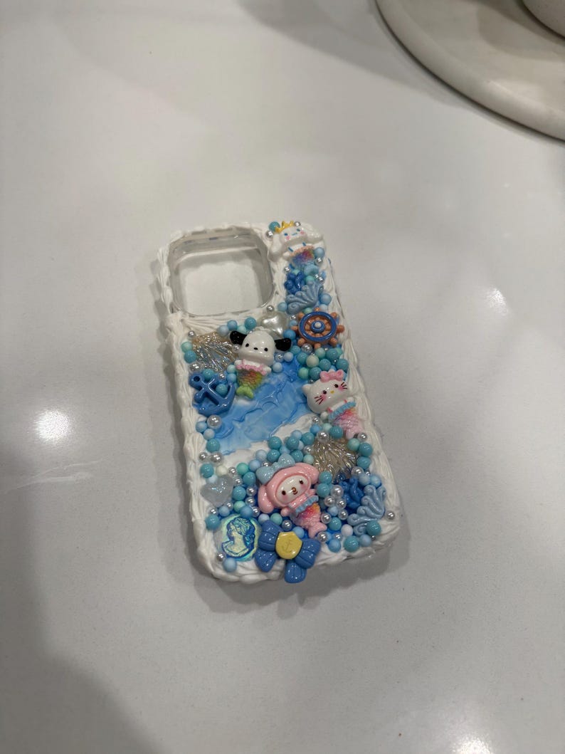 May include: A white phone case decorated with blue, pink and white 3D embellishments. The case features a mermaid theme with seashells, starfish and a small mermaid figurine.