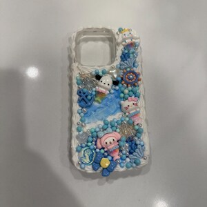 May include: A white phone case decorated with blue, pink and white 3D embellishments. The embellishments include seashells, starfish and cartoon characters.