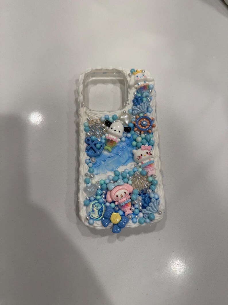 May include: A white phone case decorated with blue, pink and white 3D embellishments. The embellishments include seashells, starfish and cartoon characters.