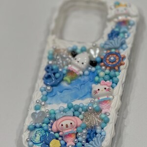 May include: A white phone case decorated with a blue and white ocean theme. The case features a mermaid-themed Hello Kitty, a mermaid-themed My Melody, and a blue anchor. The case is covered in blue and white beads, seashells, and other ocean-themed embellishments.