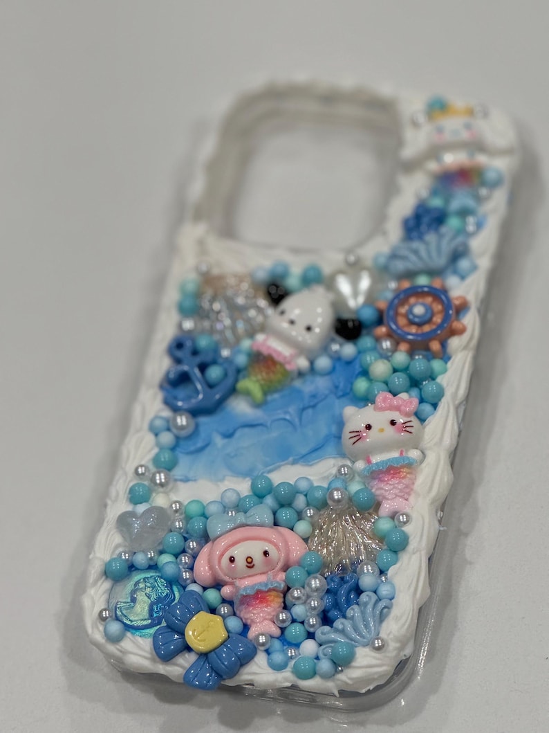 May include: A white phone case decorated with a blue and white ocean theme. The case features a mermaid-themed Hello Kitty, a mermaid-themed My Melody, and a blue anchor. The case is covered in blue and white beads, seashells, and other ocean-themed embellishments.