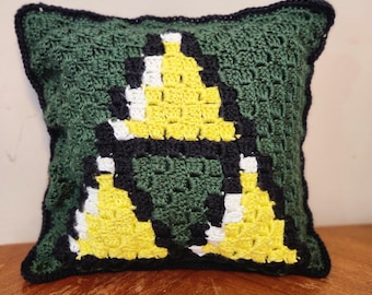 May include: A handmade crochet pillow featuring a pixelated design in shades of green, yellow, black, and white. The pillow has a dark green background with a black border. The central design is a yellow triangle with black and white accents.