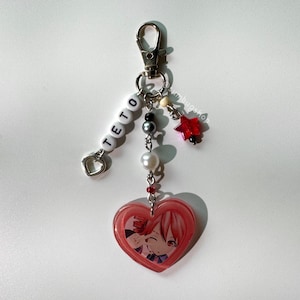 May include: A keychain featuring a silver lobster clasp, a strand of white letter beads spelling TETO with a heart charm, and a central strand with grey, white, and red beads. A red star charm and a pink heart-shaped pendant with an anime character.